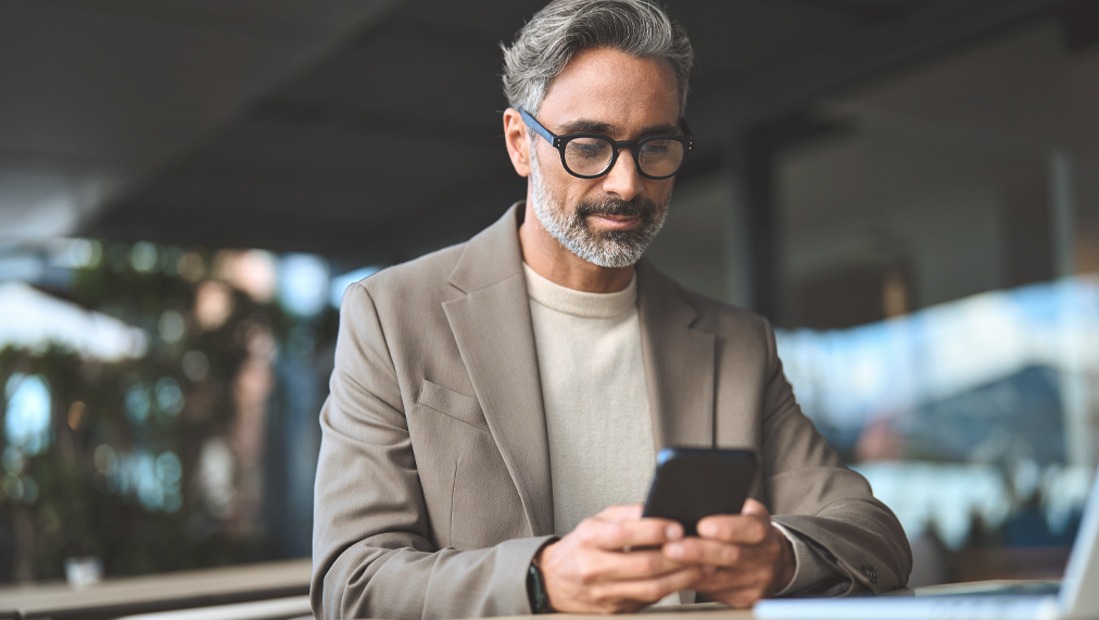 Stylish older busy businessman investor wearing glasses holding smartphone looking at cellphone doing financial payments. Happy middle aged business man using mobile phone sitting outside office.